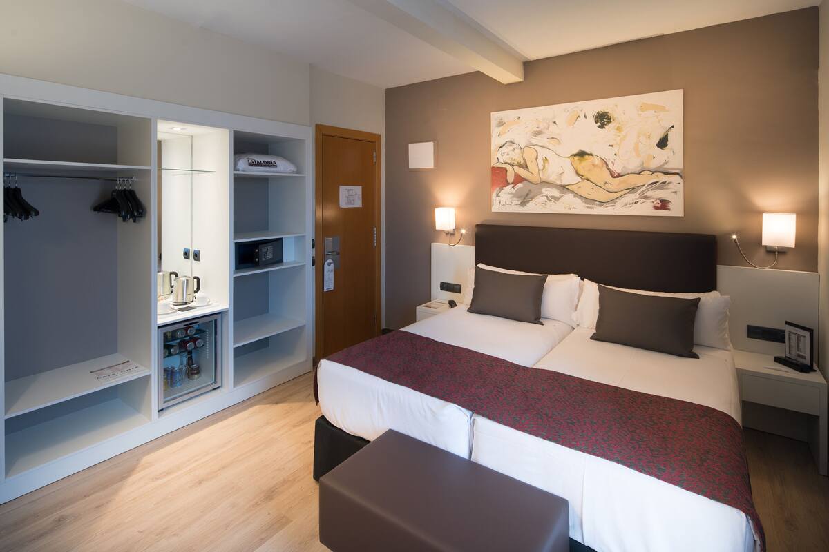 A hotel room with a large bed, a wardrobe with hanging clothes, a small seating area, and a painting on the wall.