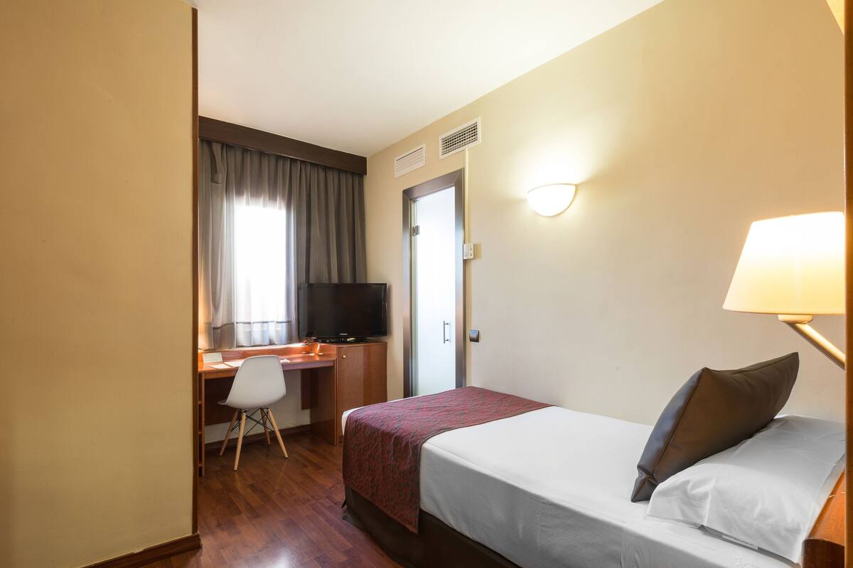 A hotel room with a bed, a desk with a chair, a television, and a door.