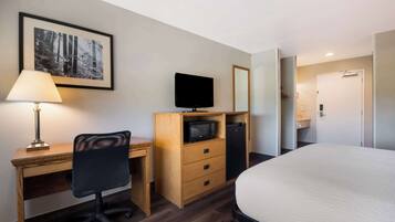 A hotel room with a wooden desk, a black office chair, a television, a microwave, a refrigerator, a bed, and a framed picture on the wall.