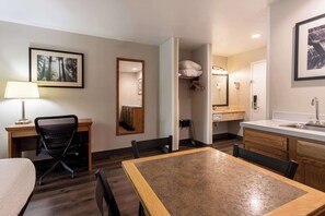 A hotel room with a wooden dining table, a desk with a chair, a bed, a bathroom with a sink and mirror, and a framed picture on the wall.