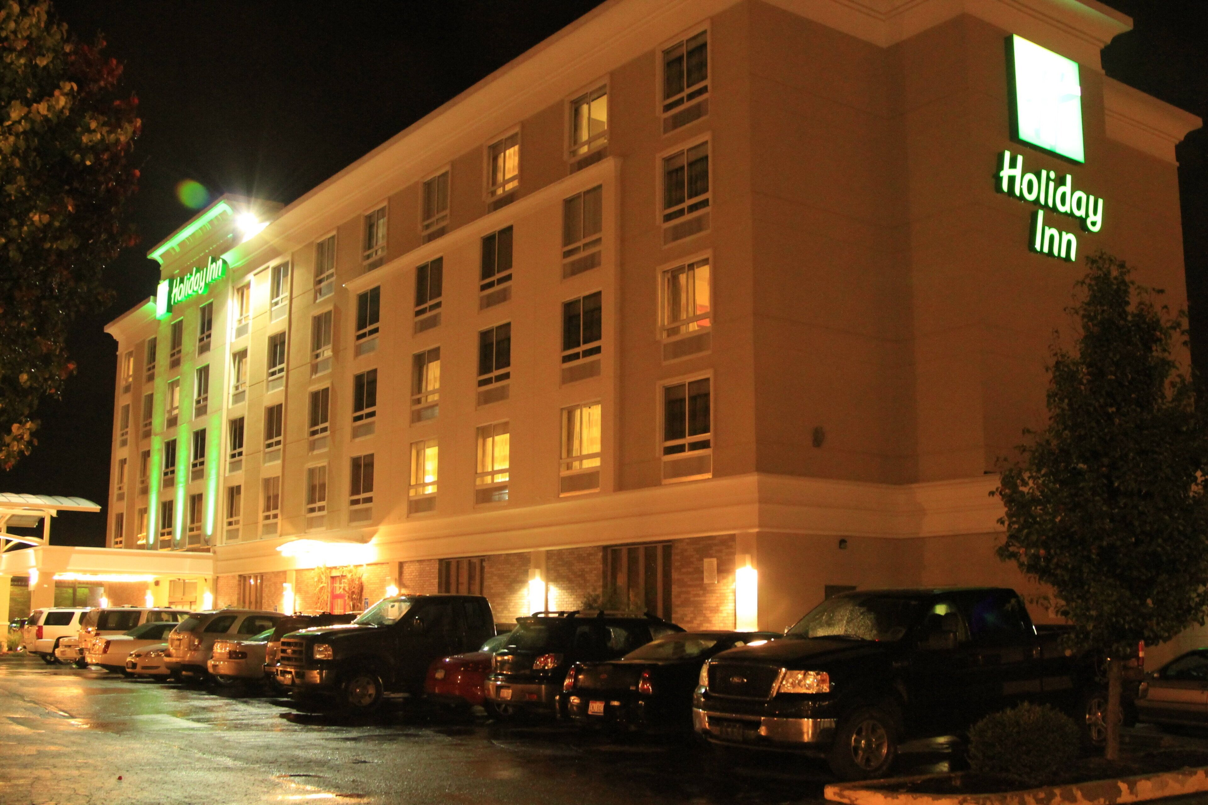 Edifício do hotel Holiday Inn com vários andares, um estacionamento com diversos carros e uma entrada bem iluminada.