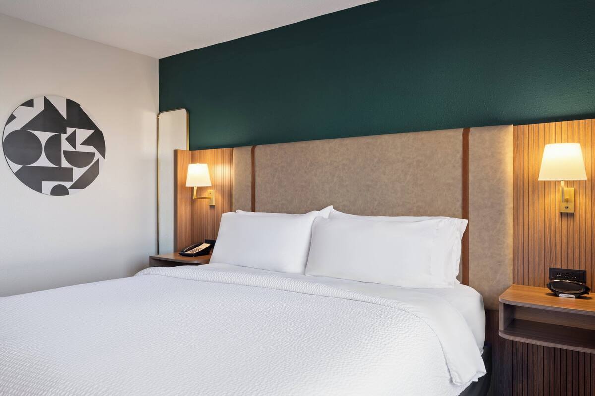 Ein Hotelzimmer mit einem großen Bett, zwei Nachttischen inklusive Lampen, einem Wandtelefon und einem geometrischen Wandbild.