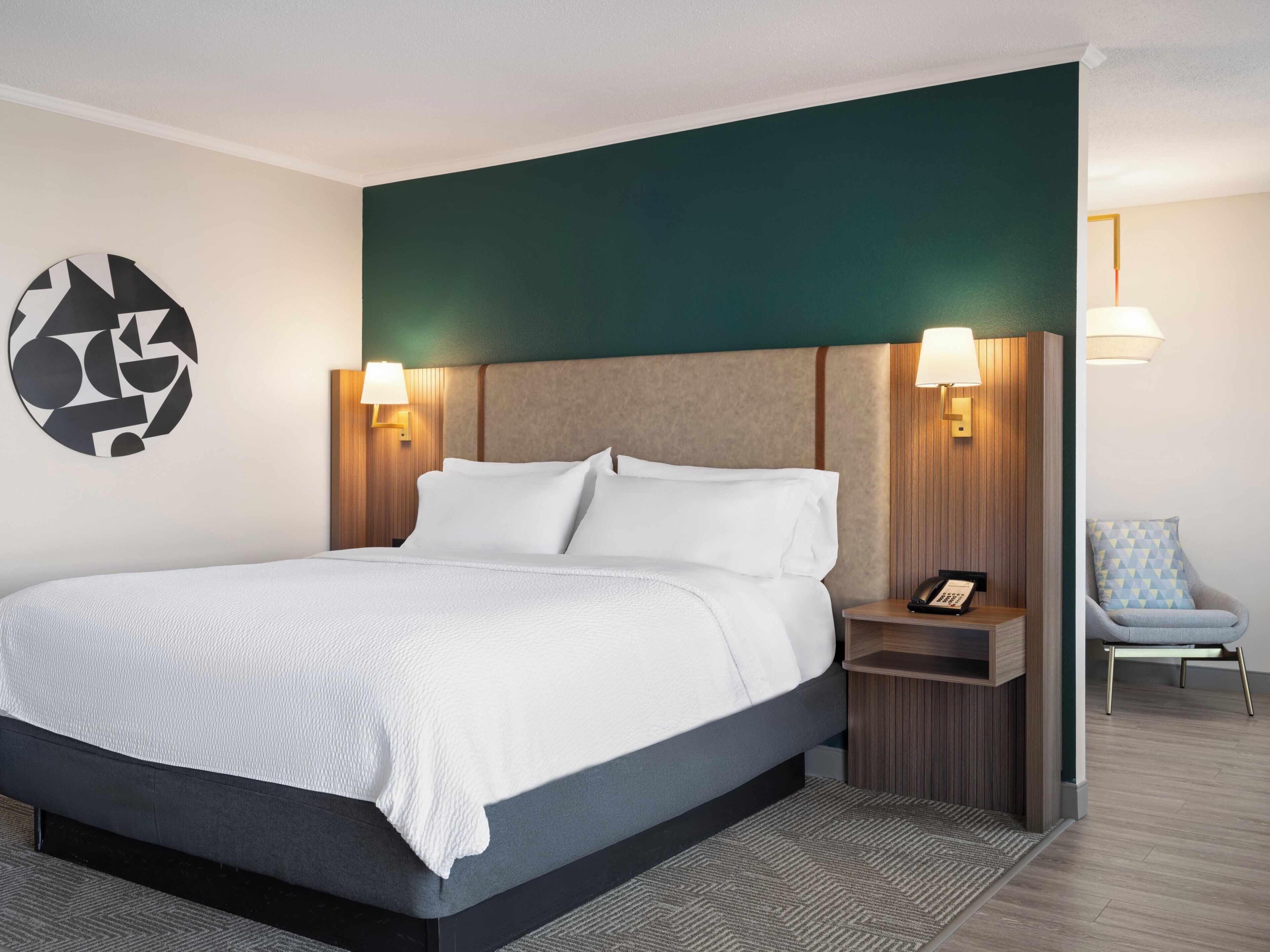 Una habitación de hotel moderna con una cama grande, dos lámparas de noche, una silla y una obra de arte en la pared.