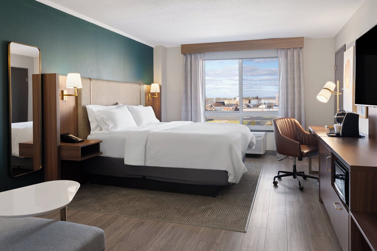 A hotel room with a large bed, a desk with a chair, a TV, and a window with a city view.