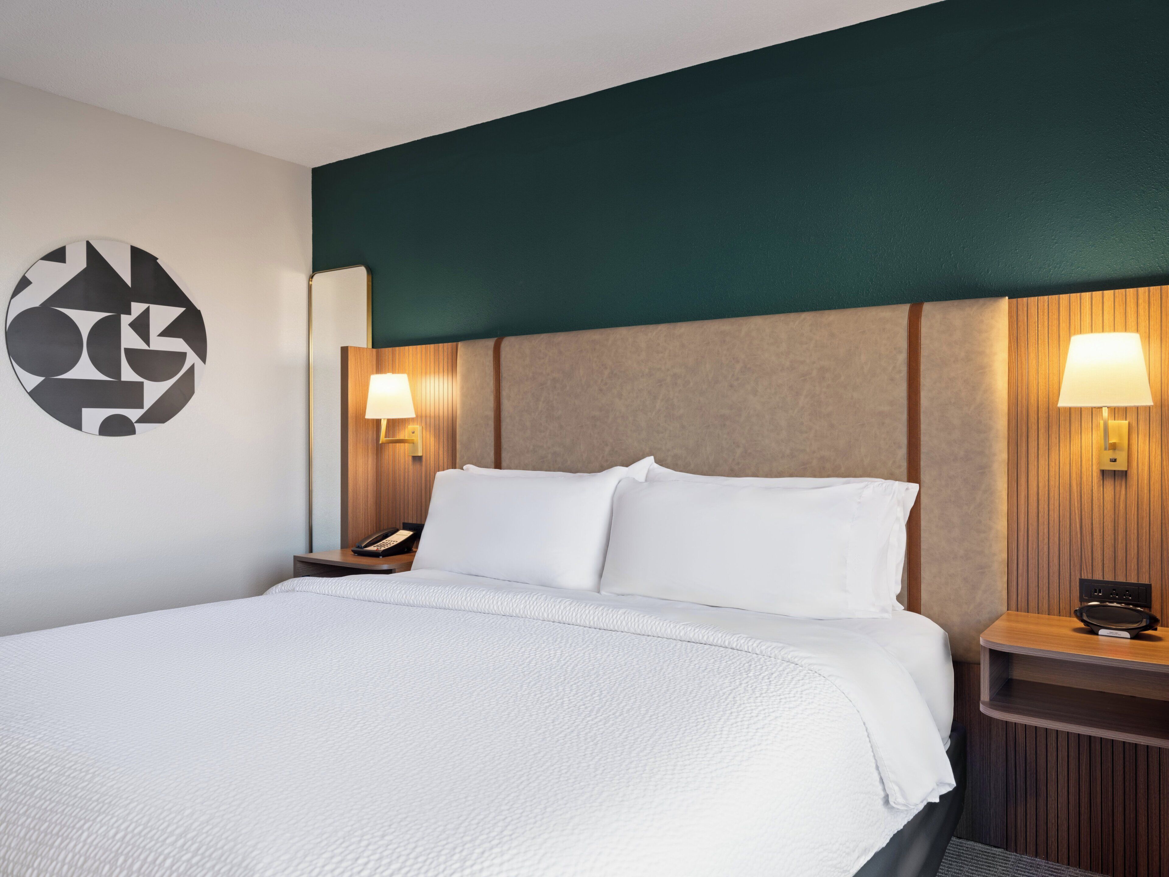 Hotelkamer met een groot bed, twee nachtkastjes met lampen, een telefoon aan de muur en een geometrisch wanddecoratie.
