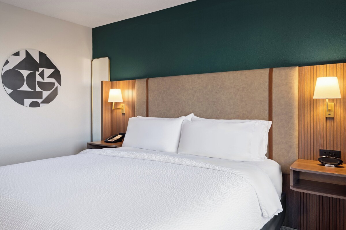 A hotel room with a large bed, two bedside tables with lamps, a wall-mounted phone, and a geometric wall decoration.