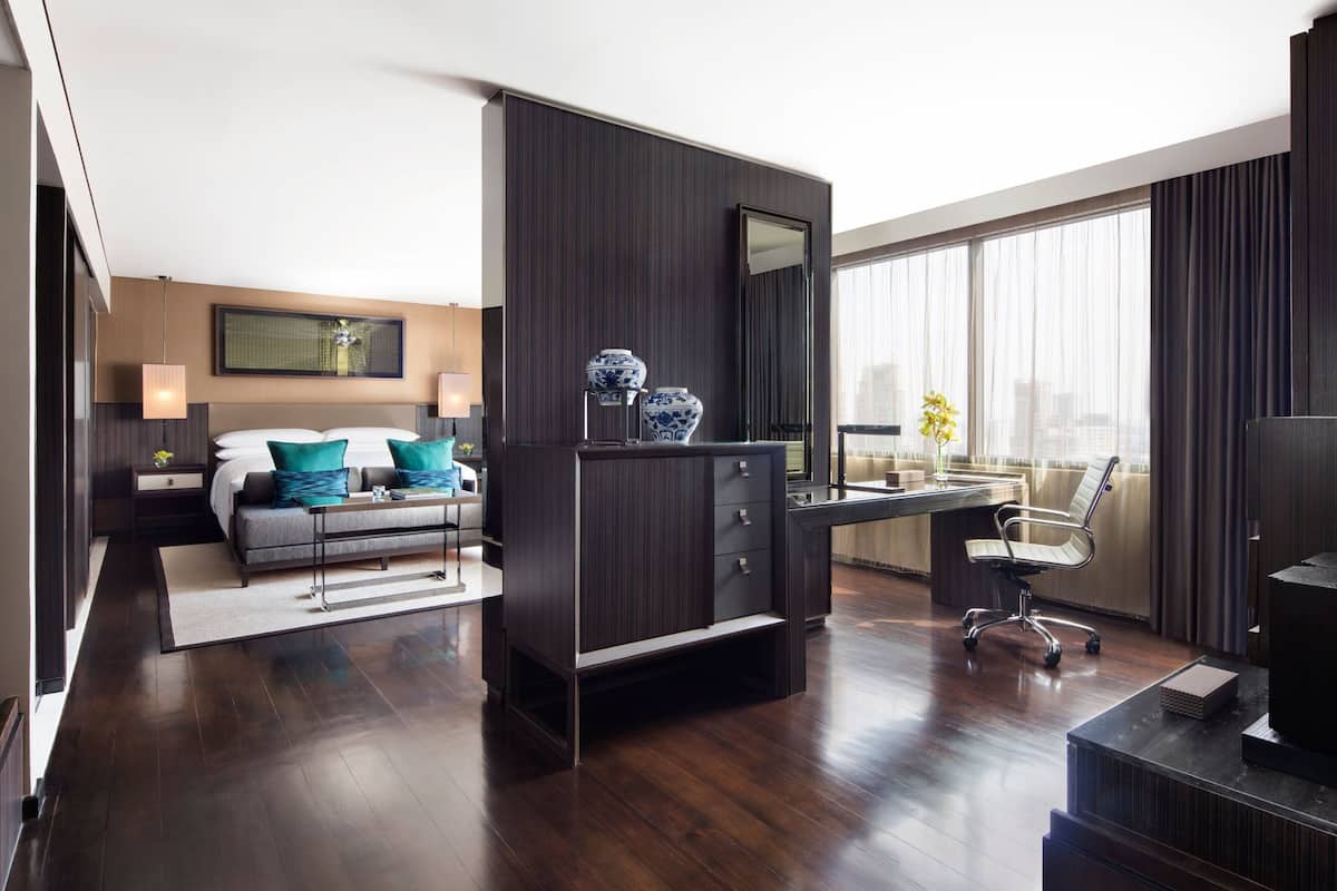 A modern hotel room with a dark wood floor, a bed with teal pillows, a desk with a chair, a large window with curtains, and a city view.