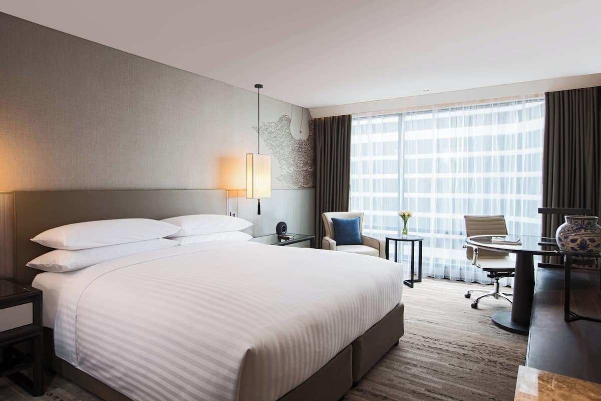 A hotel room with a large bed, a desk, a chair, a lamp, and a view of the city through the window.