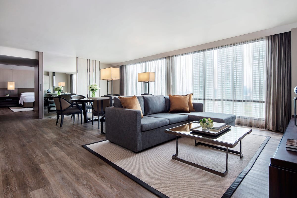 A modern hotel room with a grey sofa, a dining table with chairs, a coffee table, and large windows with curtains.