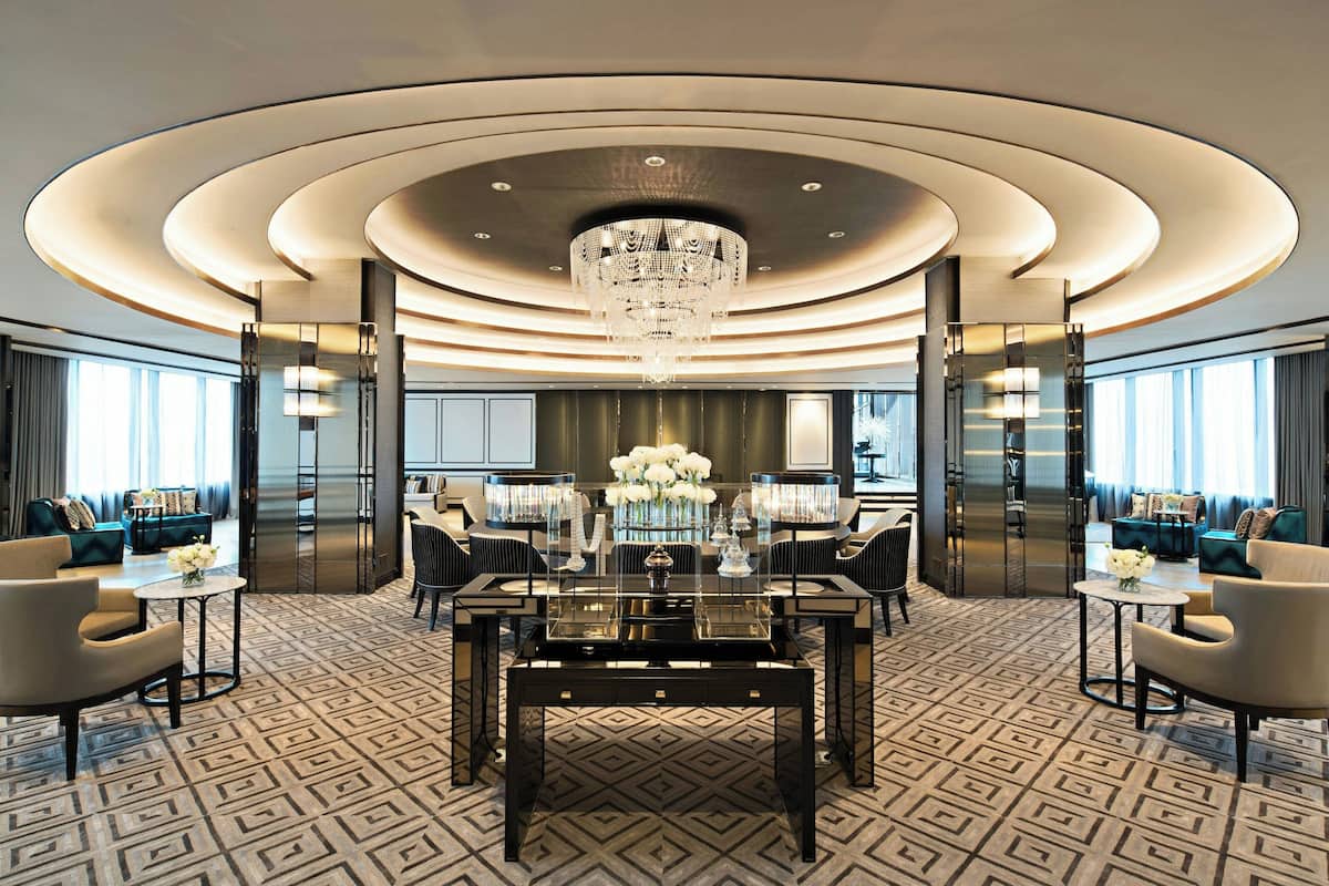 A luxurious hotel lobby with a central round table, elegant chandeliers, and modern seating arrangements.