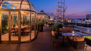 A glass-enclosed dining area with tables and chairs, overlooking a marina with boats and a city skyline at dusk. - Bangkok Marriott Marquis Queen’s Park (Bangkok)
