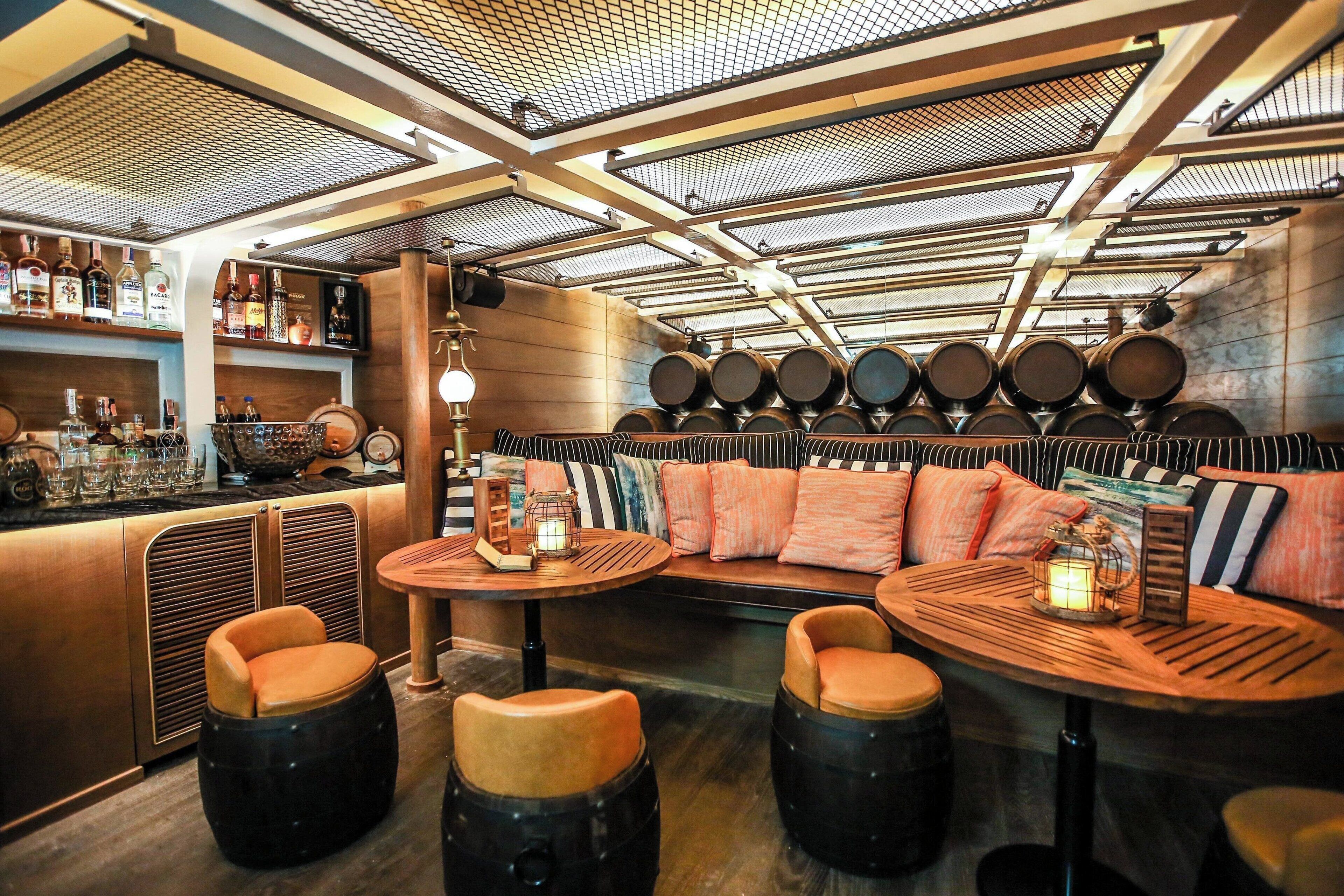 A bar area with round tables, cushioned seating, and a variety of bottles and glasses on shelves.
