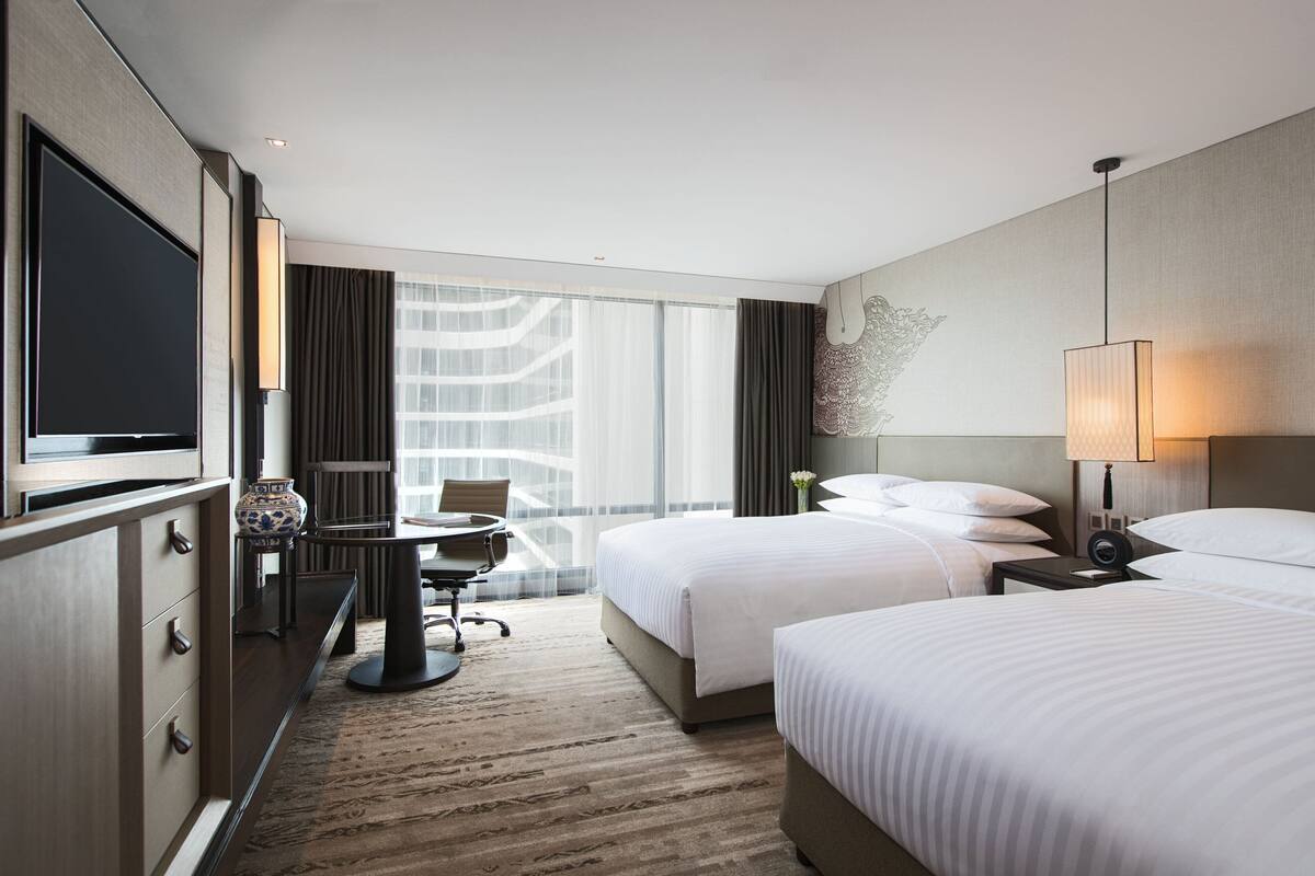 A hotel room with two beds, a desk with a chair, a TV, and a large window with curtains.