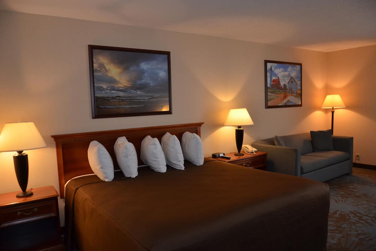 Une chambre d’hôtel avec un grand lit, deux tables de chevet avec des lampes, un fauteuil gris et deux tableaux accrochés aux murs.