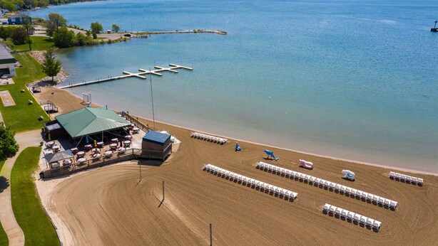 Vue aérienne d’une propriété au bord du lac, avec un quai, des chaises de plage et un espace abrité comprenant des tables et des parasols.