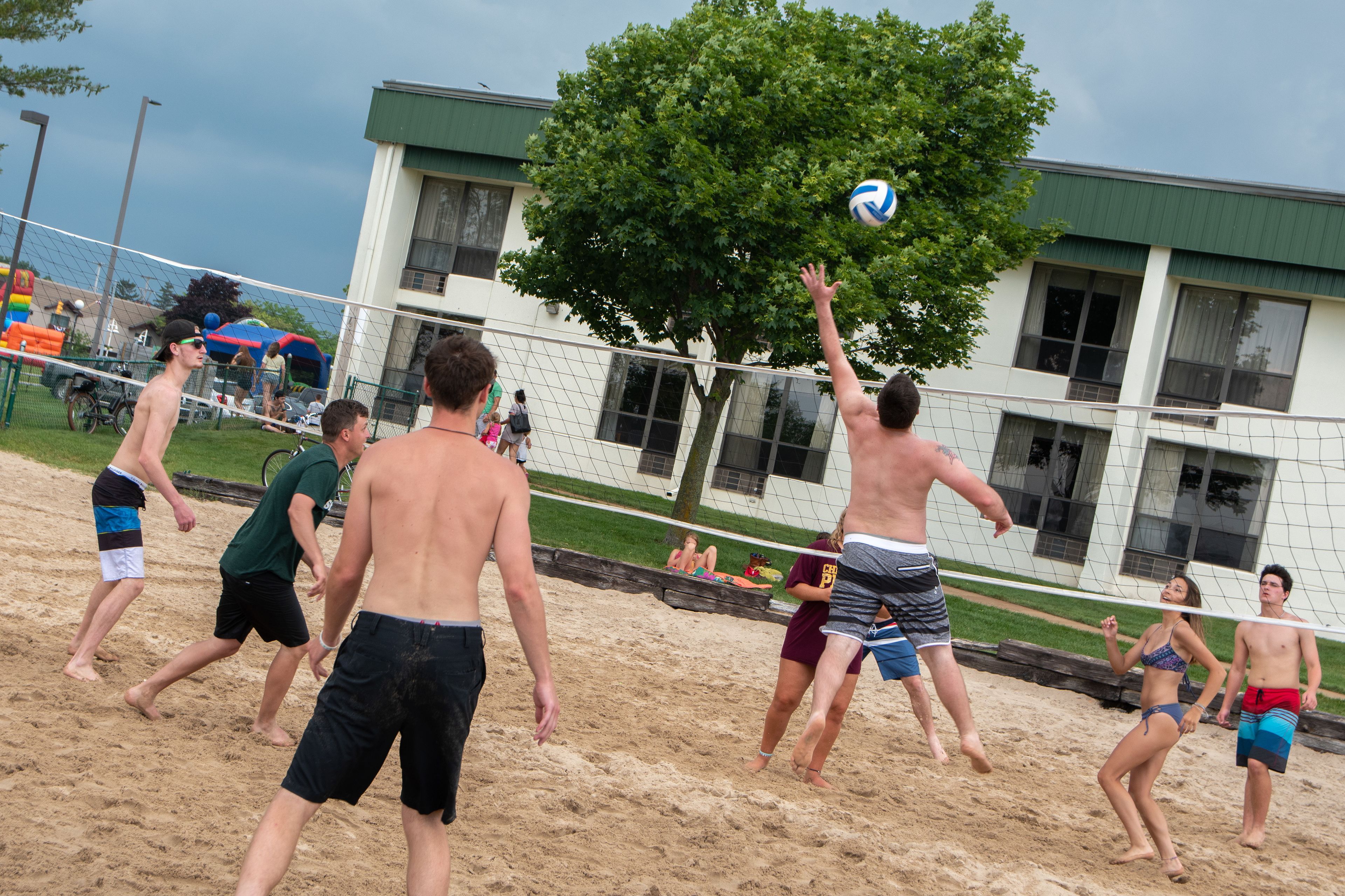 A sandy beach volleyball court with players in motion, a tree, and a building in the background.