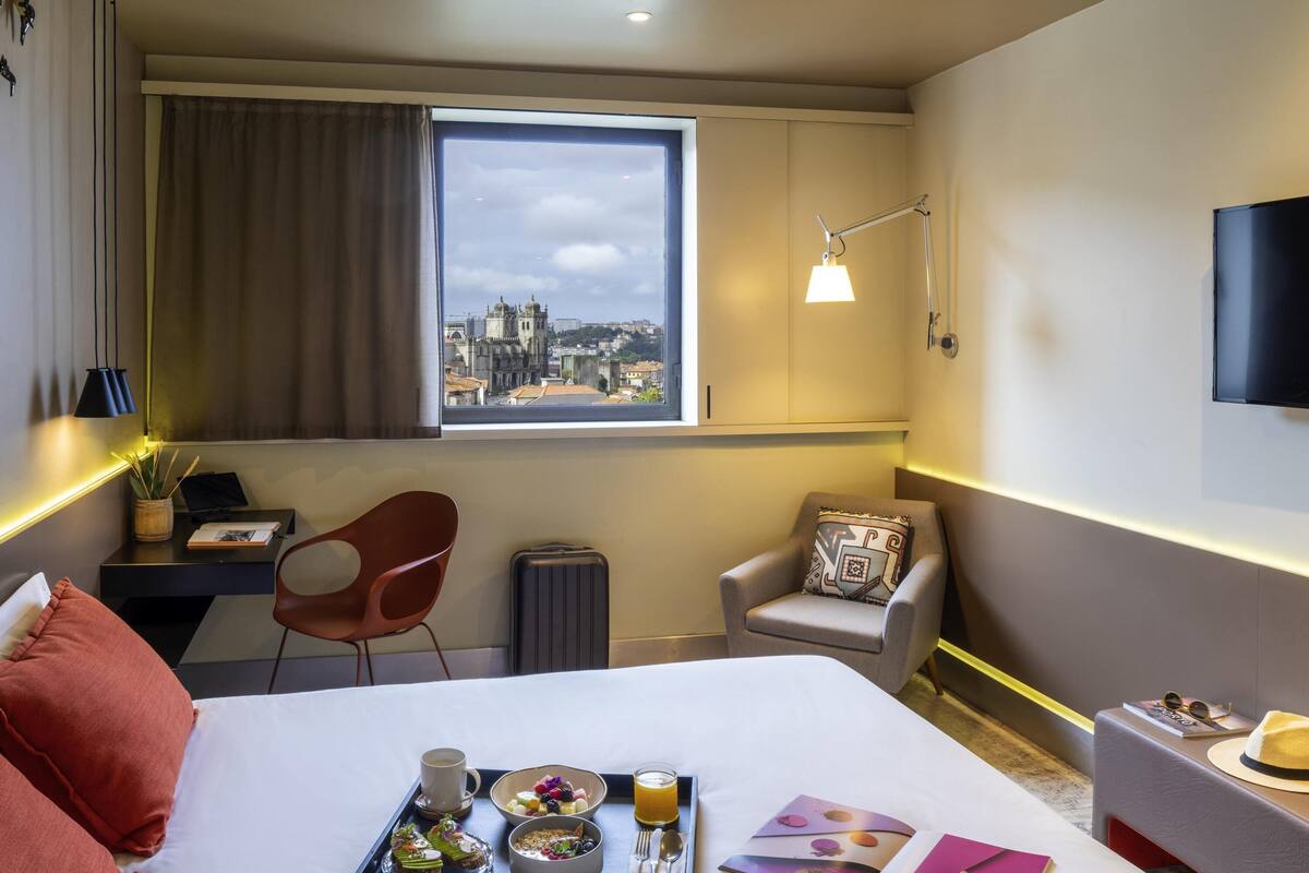 Ein Hotelzimmer mit Bett, Schreibtisch, Stuhl, einem Fenster mit Blick auf eine Stadtlandschaft und einem Tablett mit Frühstücksgebäck.