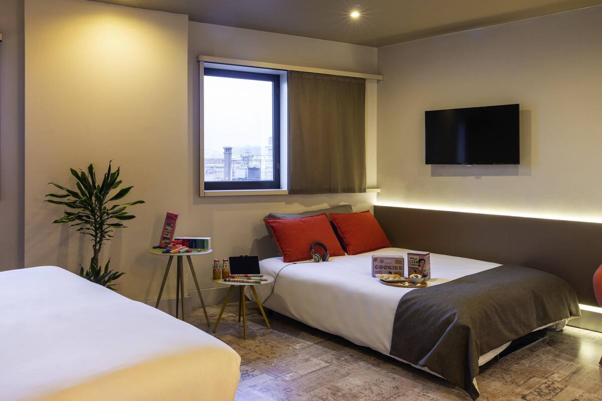 Ein Hotelzimmer mit Bett, Nachttisch, einer kleinen Pflanze, einem Fenster mit Vorhängen, einem wandmontierten Flachbildfernseher und Blick auf die Stadt.