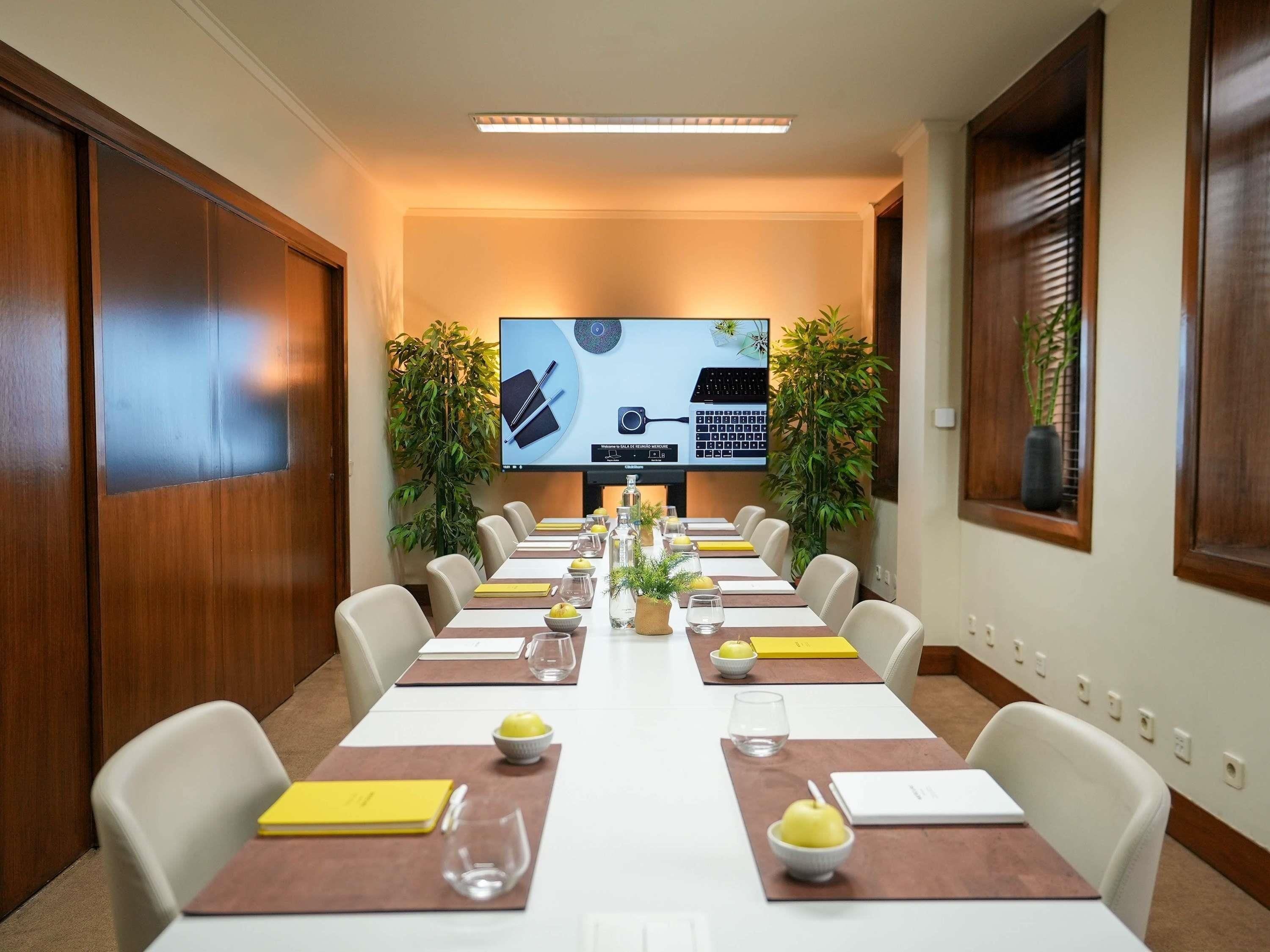 Ein Konferenzraum mit einem langen Tisch, Stühlen, einer großen Leinwand und Topfpflanzen.
