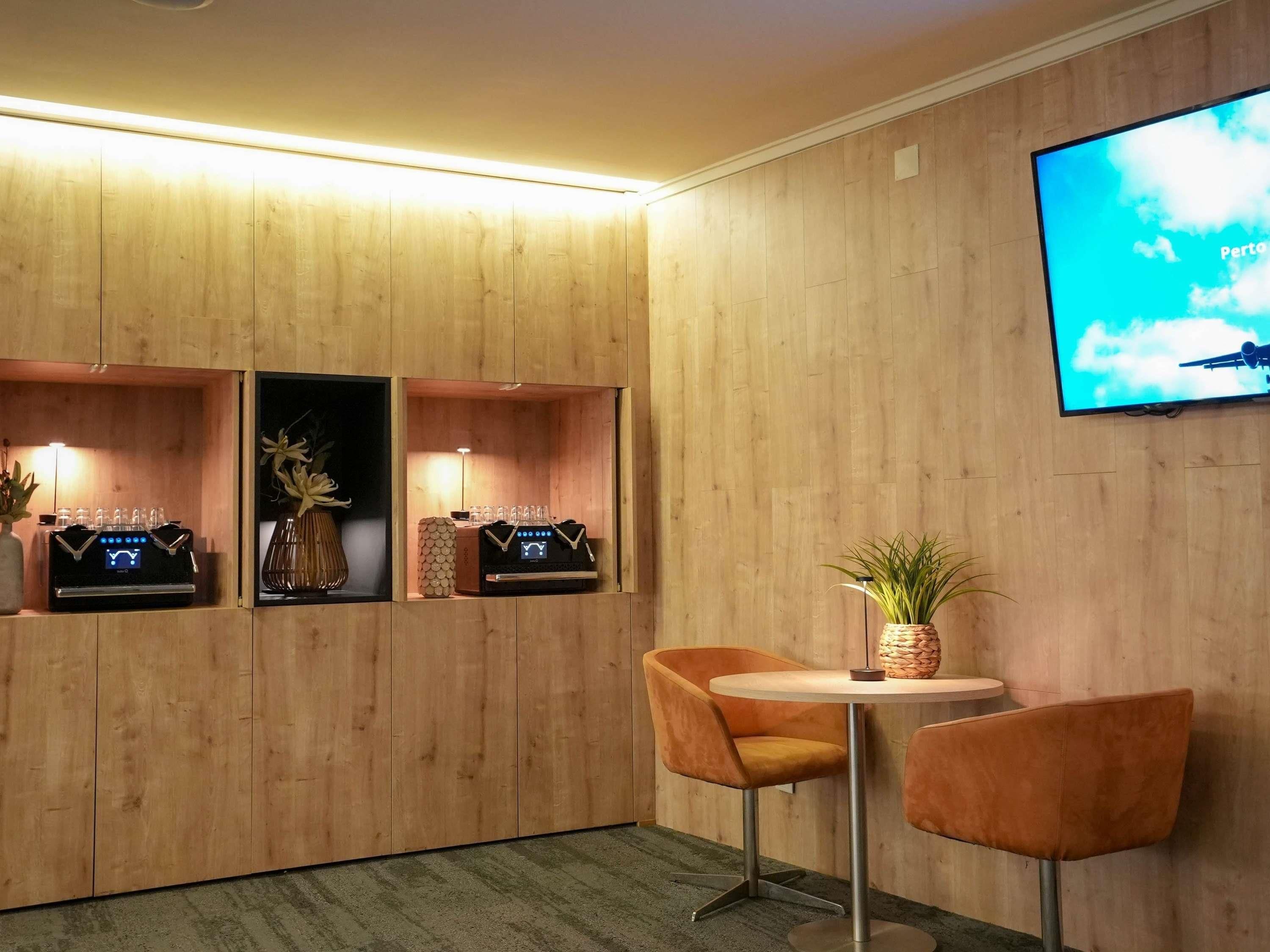 Una habitación con paneles de madera, un televisor montado en la pared, una pequeña mesa redonda con dos sillas y un dispositivo digital en un estante.