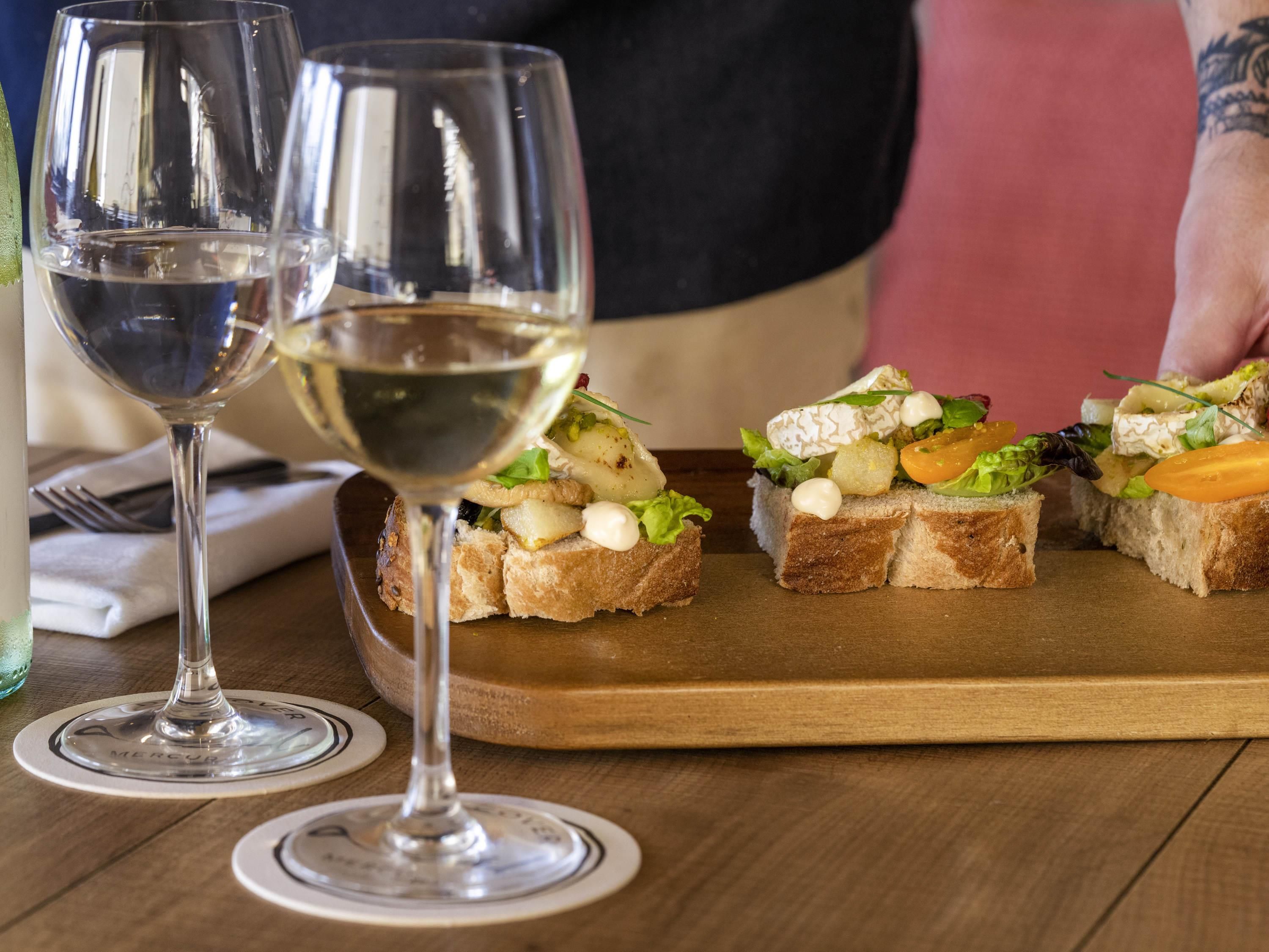 Et træbord med to glas hvidvin og en træplanke med tre stykker ristet brød toppet med forskellige ingredienser.