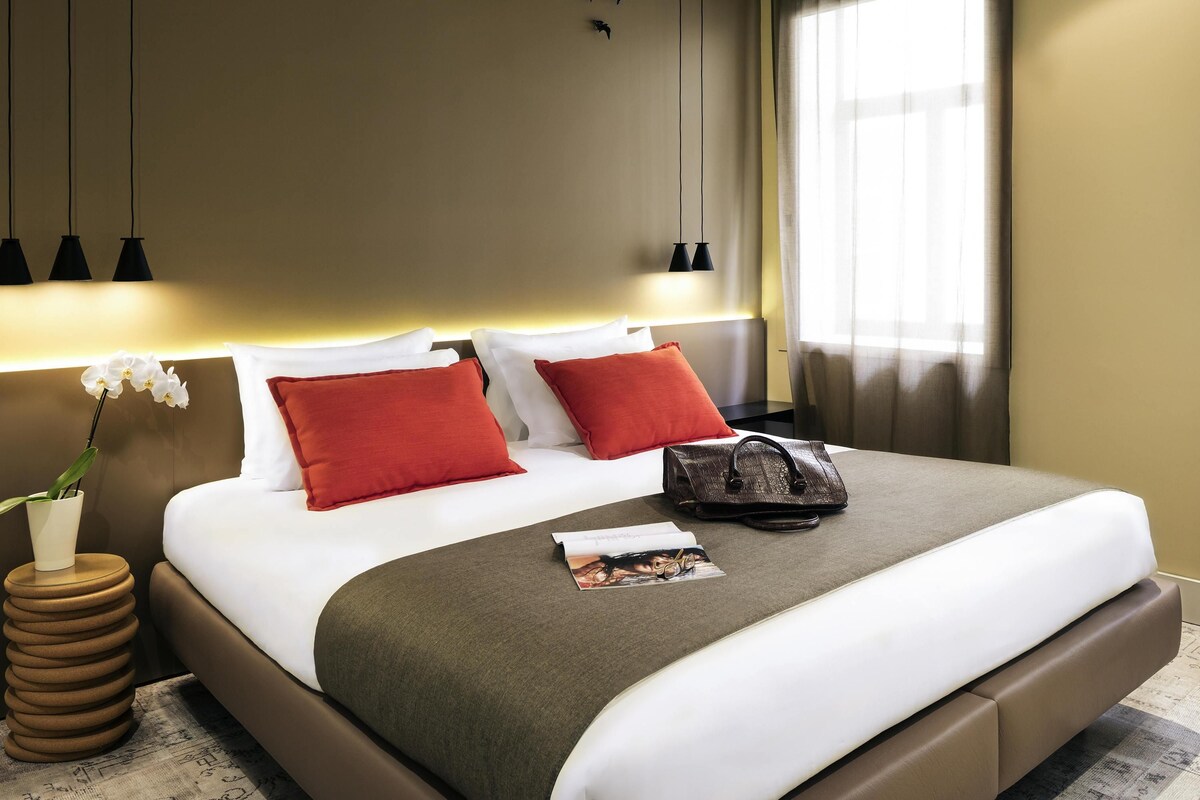 A hotel room with a bed, pillows, a nightstand, a lamp, a magazine, and a handbag.
