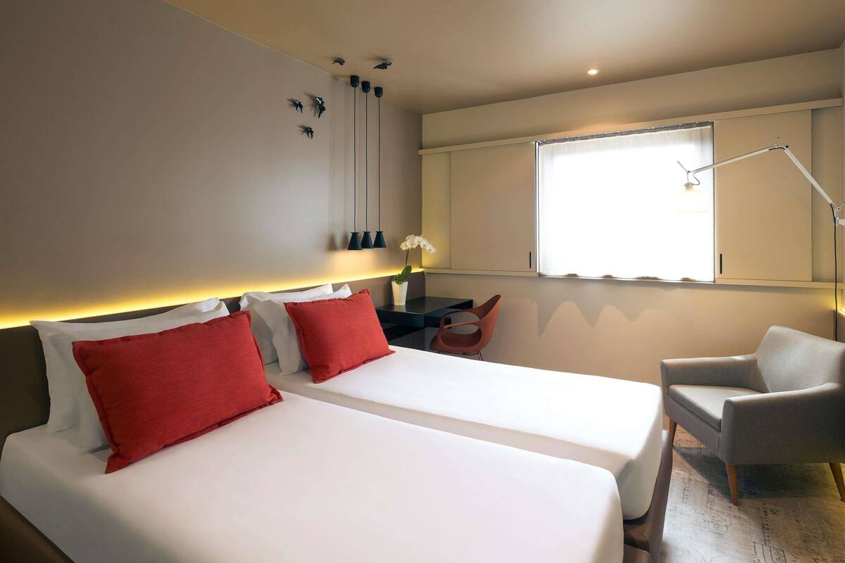 Ein Hotelzimmer mit zwei Betten, einem Schreibtisch, einem Stuhl, einem Fenster und einer Lampe.