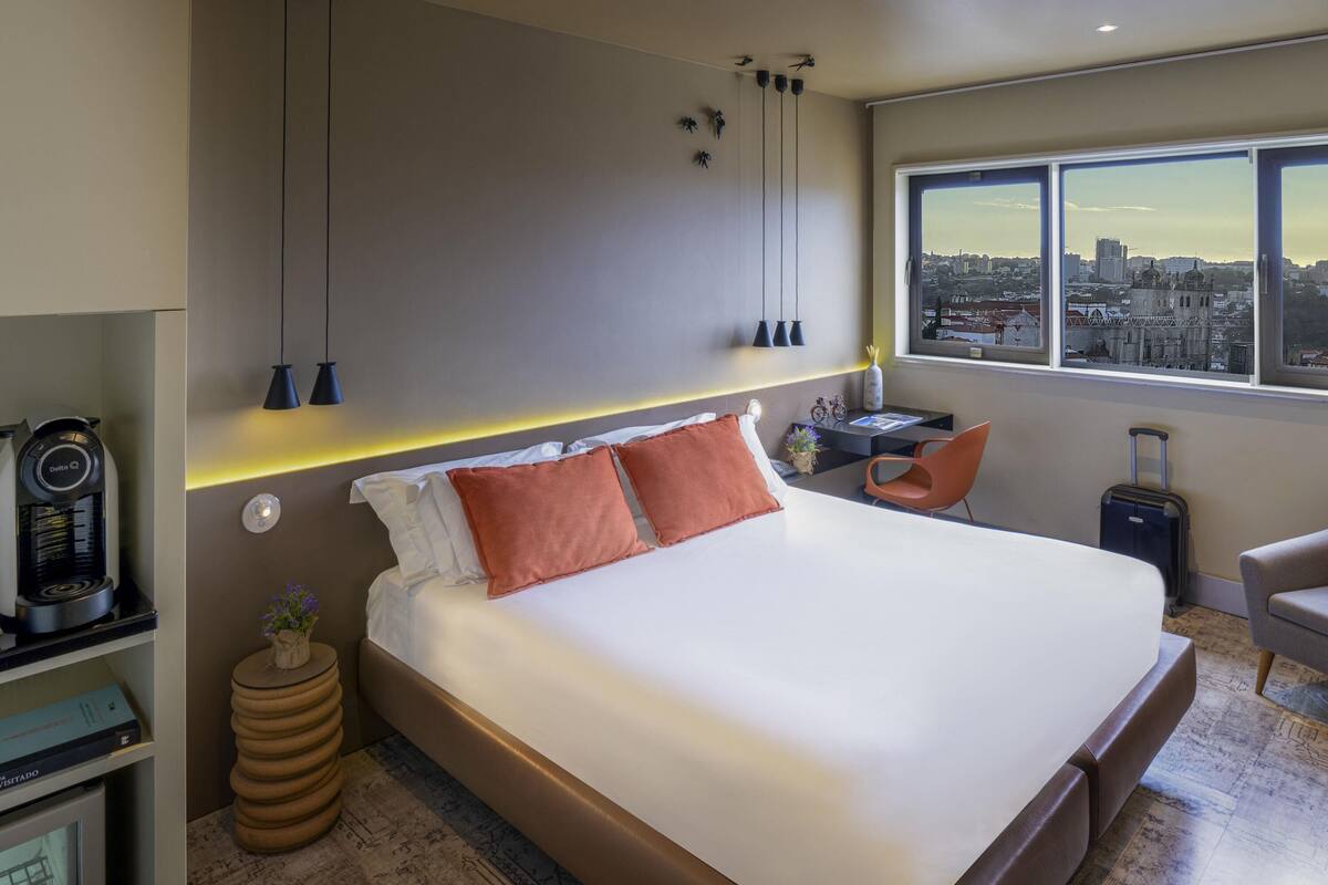Ein modernes Hotelzimmer mit einem großen Bett, einem Schreibtisch mit Stuhl, einer Kaffeemaschine, Blick auf die Stadt und einem Koffer.