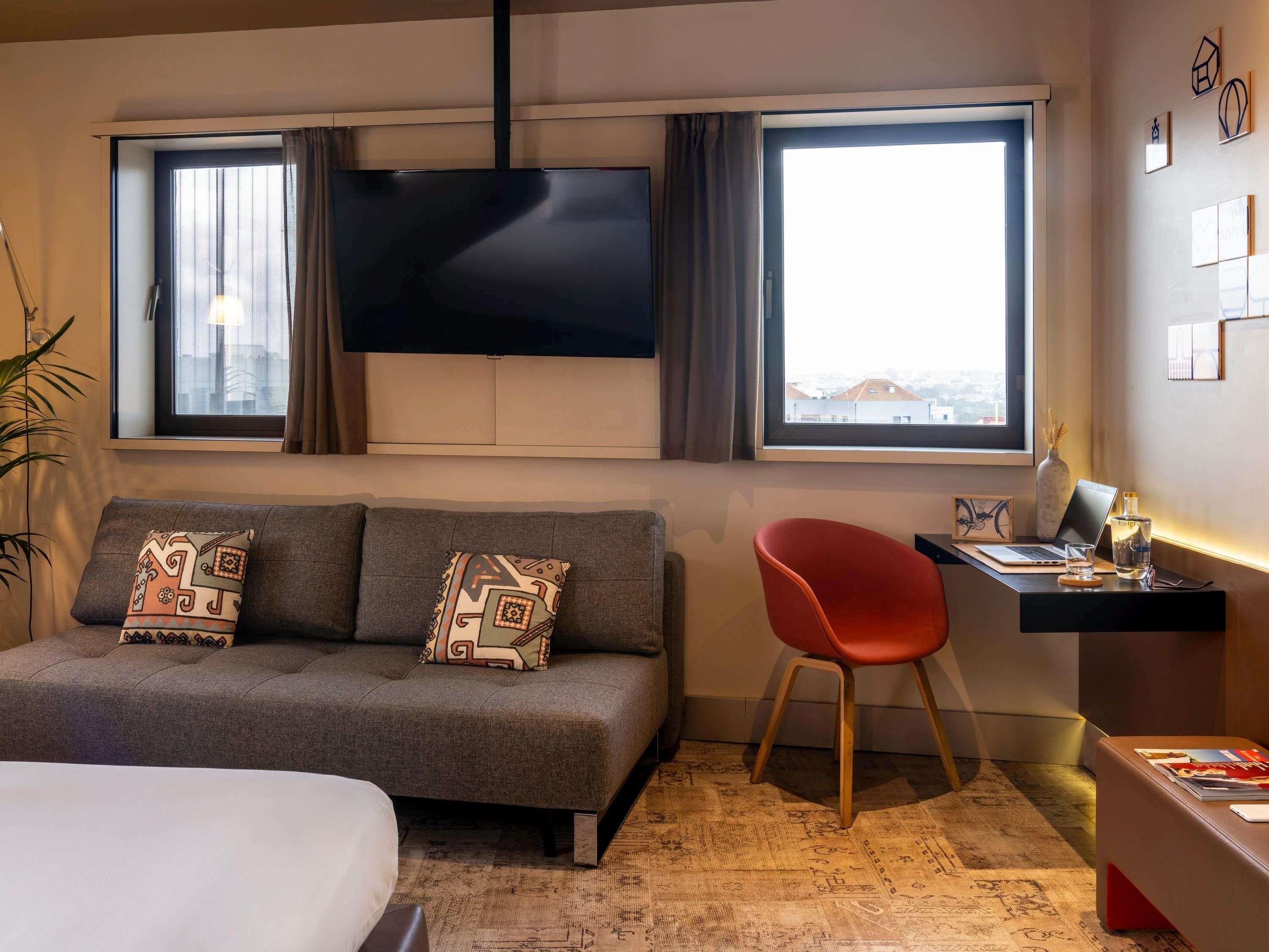 A hotel room with a sofa, a red chair, a desk with a laptop, a TV, and a view of buildings through the windows.