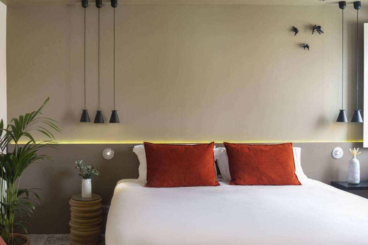 Ein modernes Hotelzimmer mit einem großen Bett, zwei roten Kissen, einer Topfpflanze und Pendelleuchten.