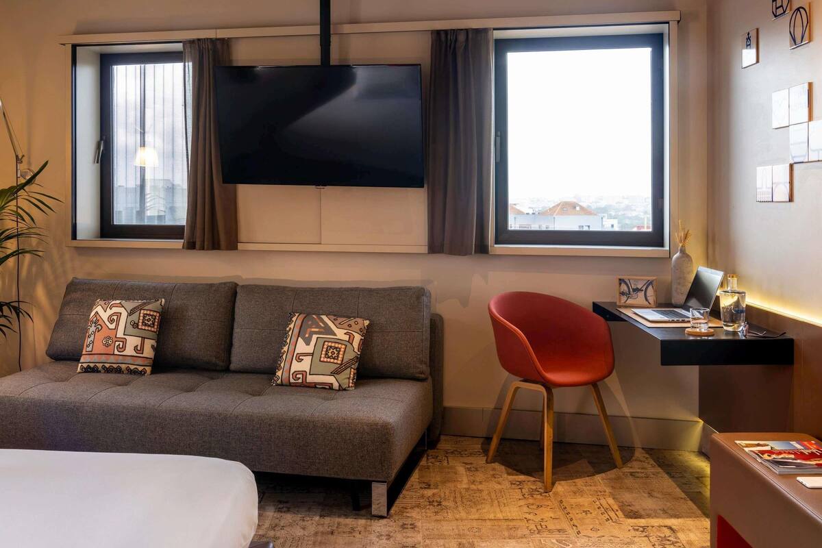 Ein Hotelzimmer mit einer Couch, einem roten Sessel, einem Schreibtisch mit einem Laptop, einem Fernseher und Blick auf Gebäude durch die Fenster.