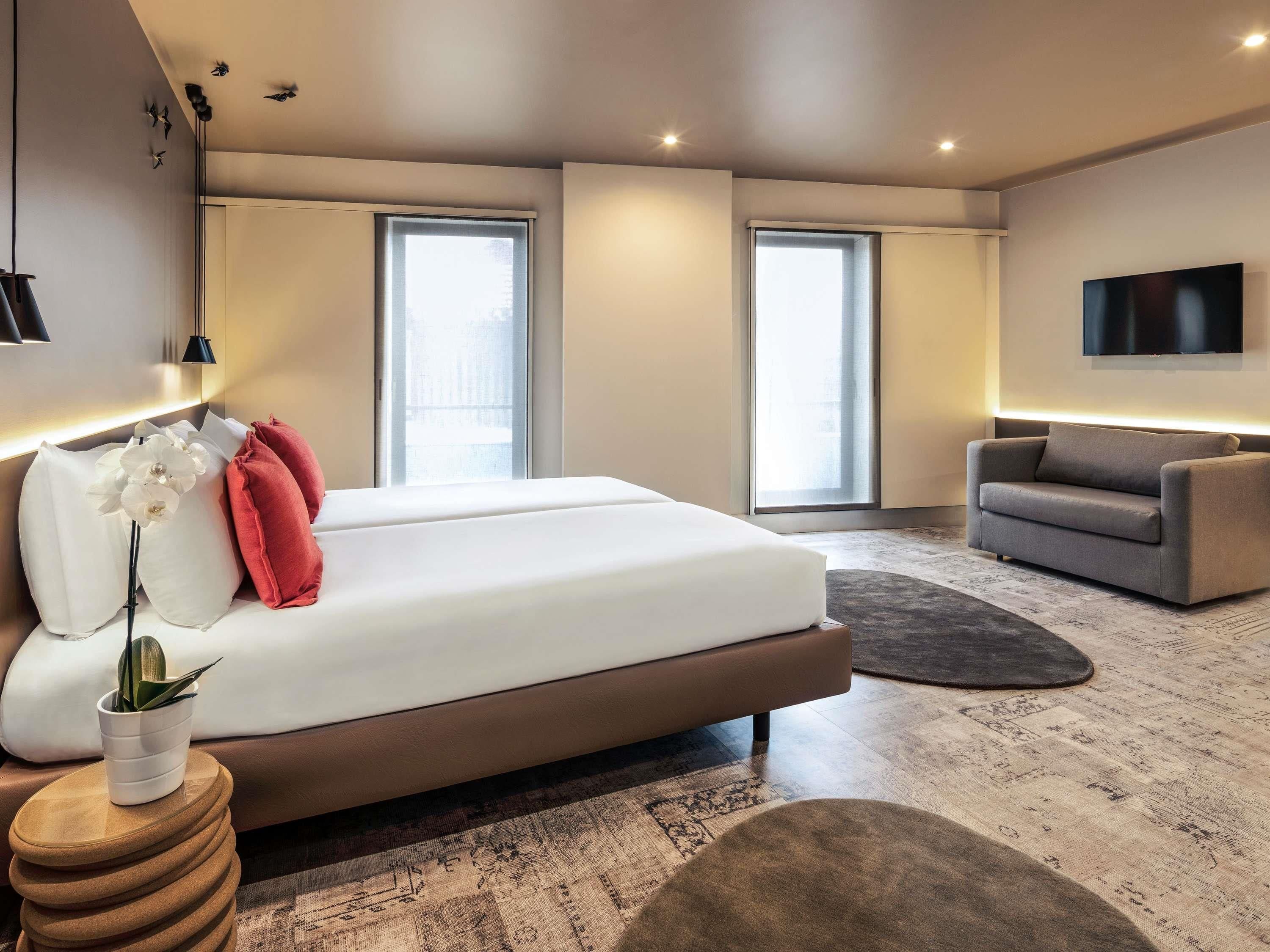 A modern bedroom with a large bed, a grey sofa, and a flat-screen TV mounted on the wall.