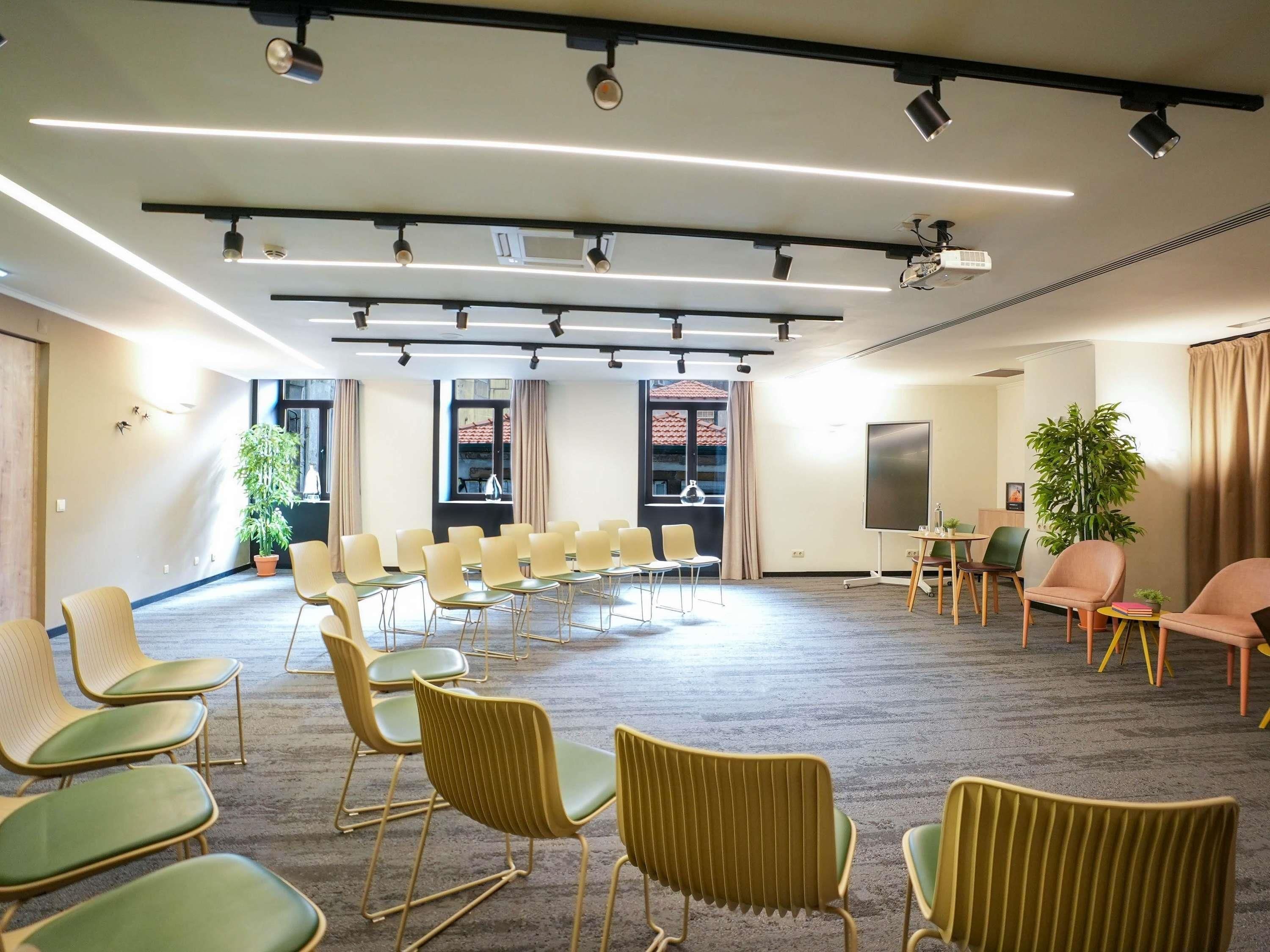 Un salón de conferencias con filas de sillas, una pantalla de proyector y un ventanal con cortinas.