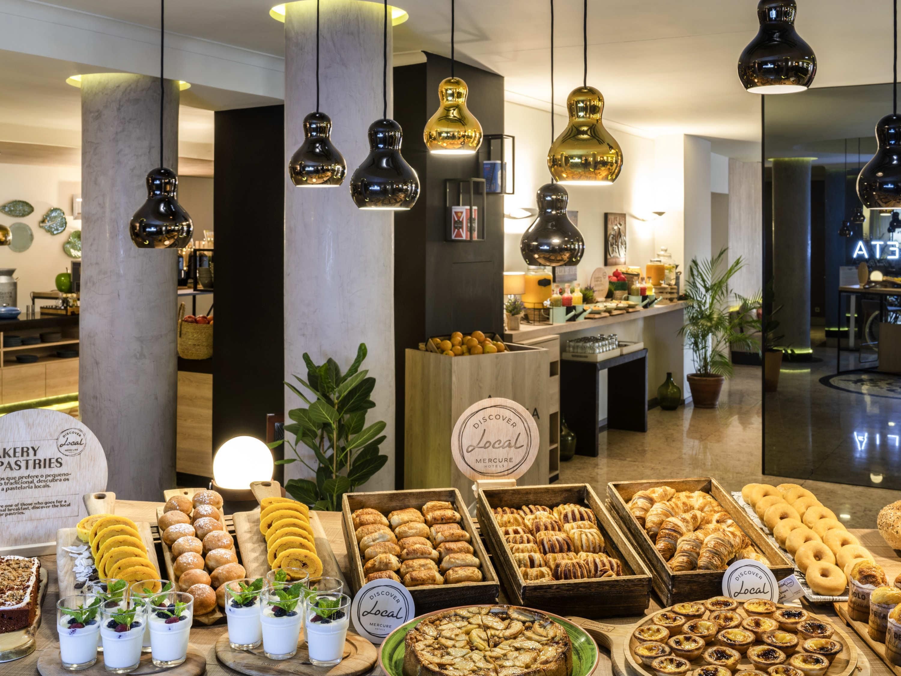 Un bufé de desayuno con diversos pasteles y bebidas, un cartel que indica 'Discover Local' y lámparas colgantes.