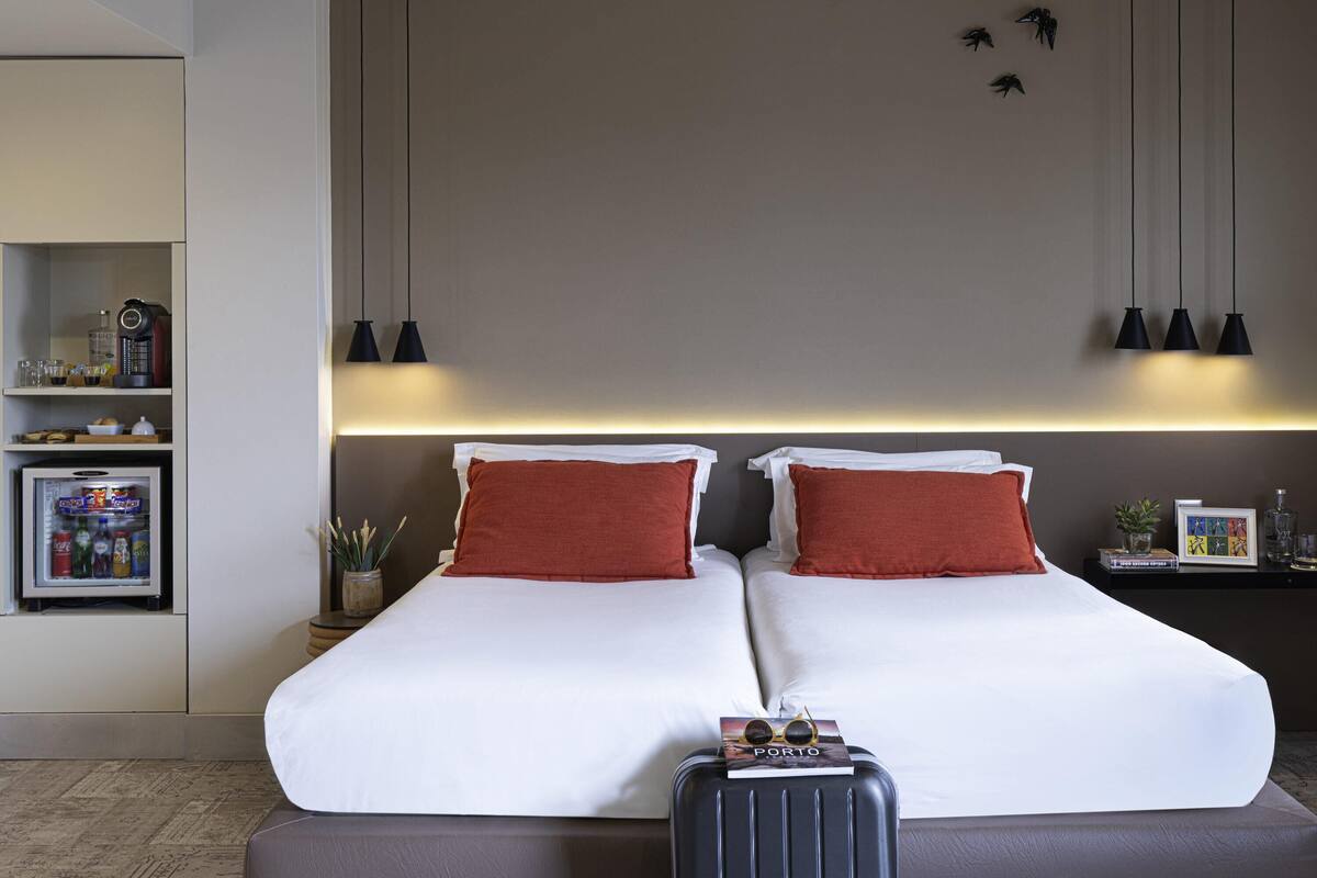 Ein Hotelzimmer mit einem Bett, einer Minibar, einem kleinen Tisch mit einer Pflanze und einem an der Wand befestigten Regal mit Gegenständen.