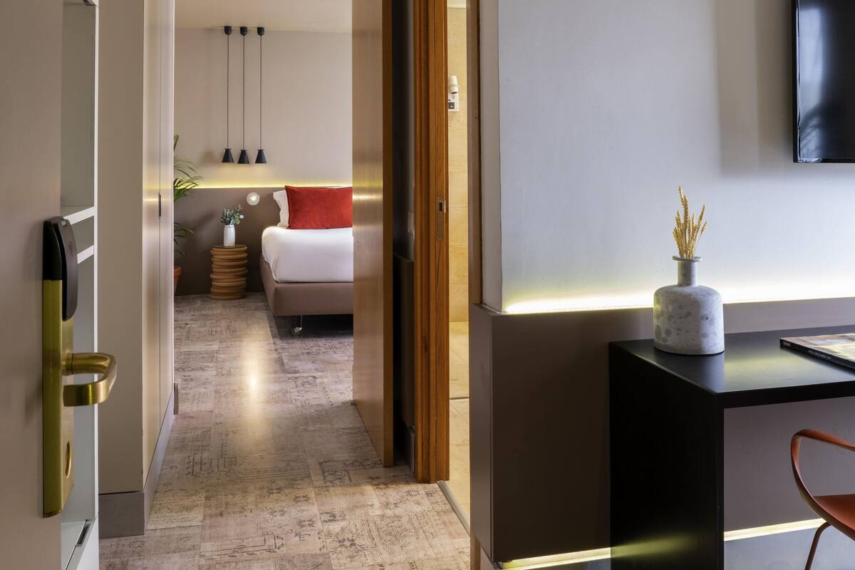 Ein modernes Hotelzimmer mit einem Bett, Nachttisch und Schreibtisch mit Stuhl. Eine Tür mit Griff, eine Wandleuchte und eine kleine Pflanze auf dem Schreibtisch sind ebenfalls vorhanden.