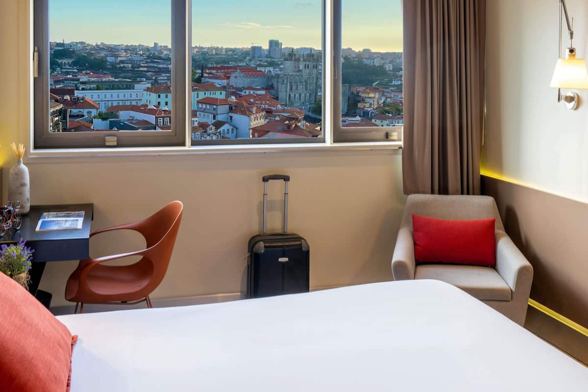 Quarto de hotel com cama, escrivaninha, cadeira, mala e vista para a cidade.