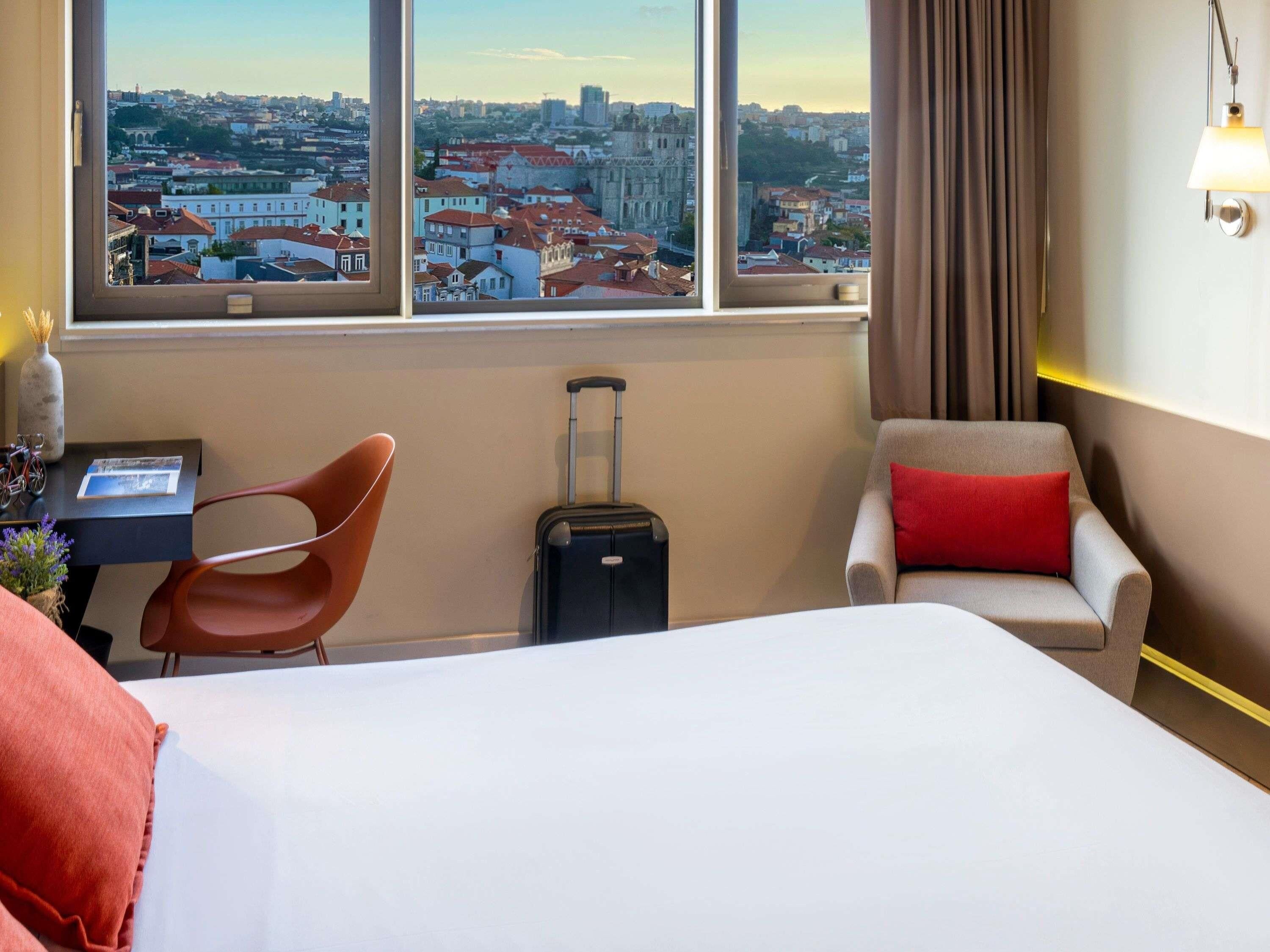Habitación de hotel con cama, escritorio, silla, maleta y vistas a la ciudad.