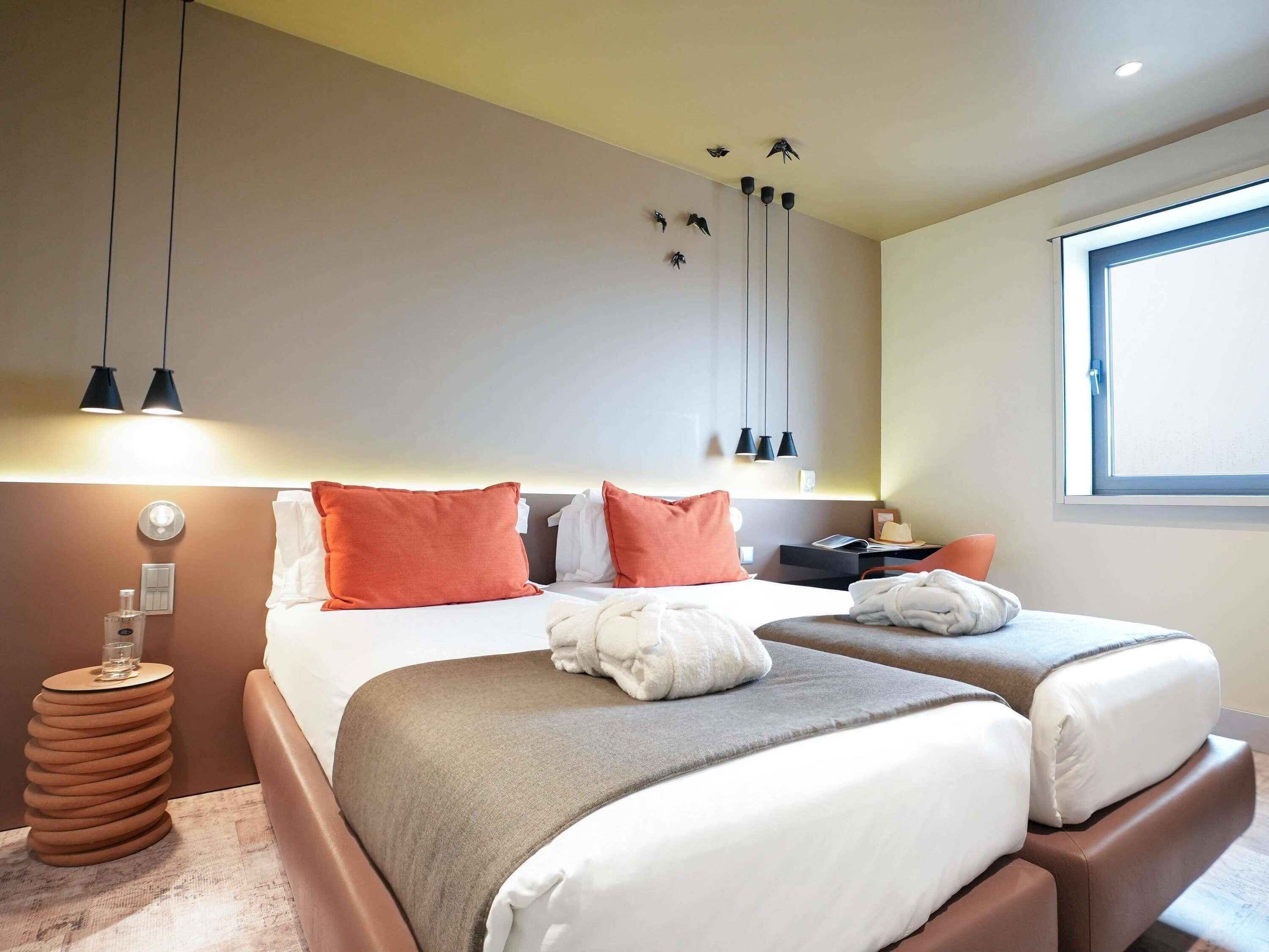 A hotel room with two beds, a bedside table, a lamp, a window, and a wall-mounted shelf.