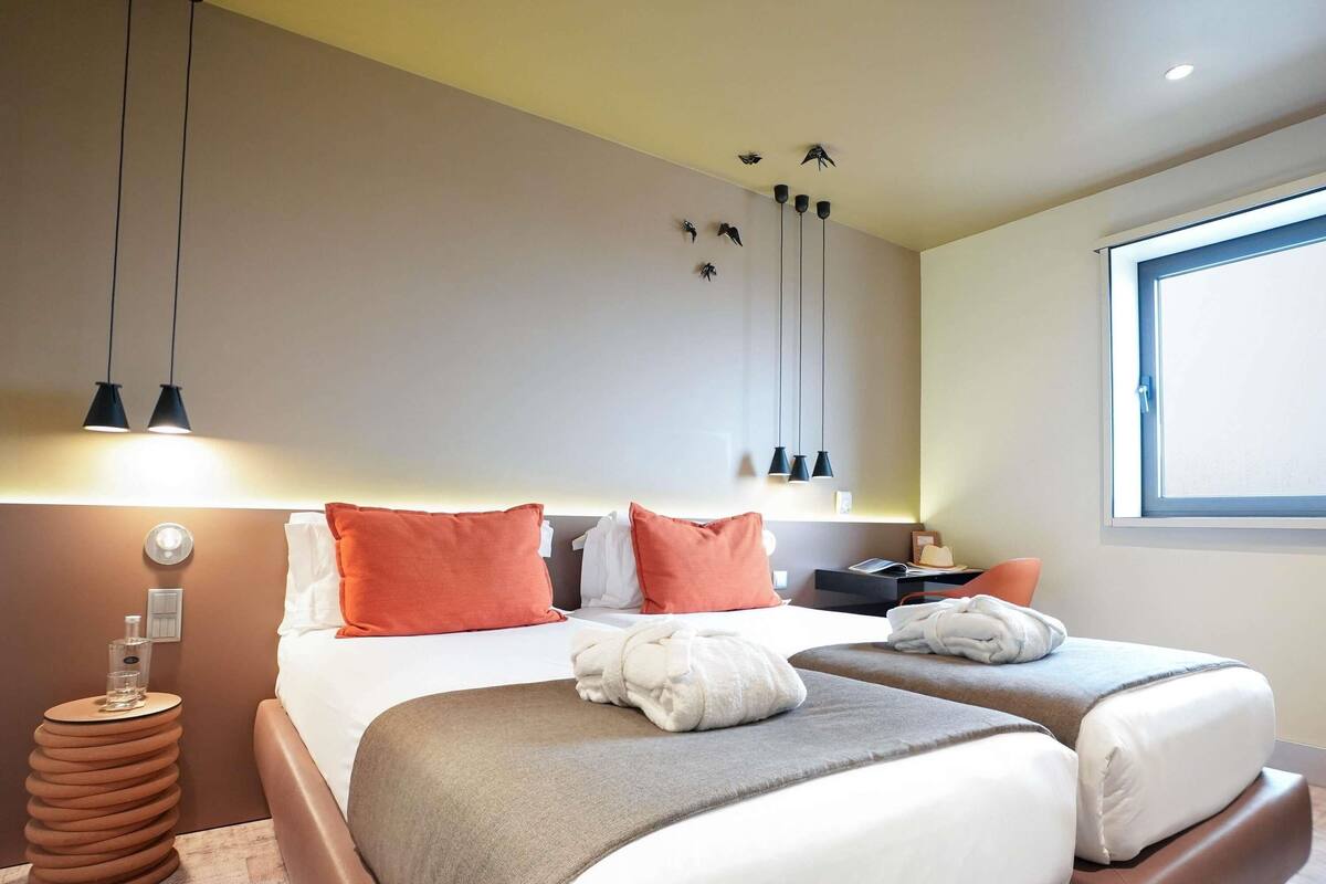 Ein Hotelzimmer mit zwei Betten, einem Nachttisch, einer Lampe, einem Fenster und einem an der Wand befestigten Regal.