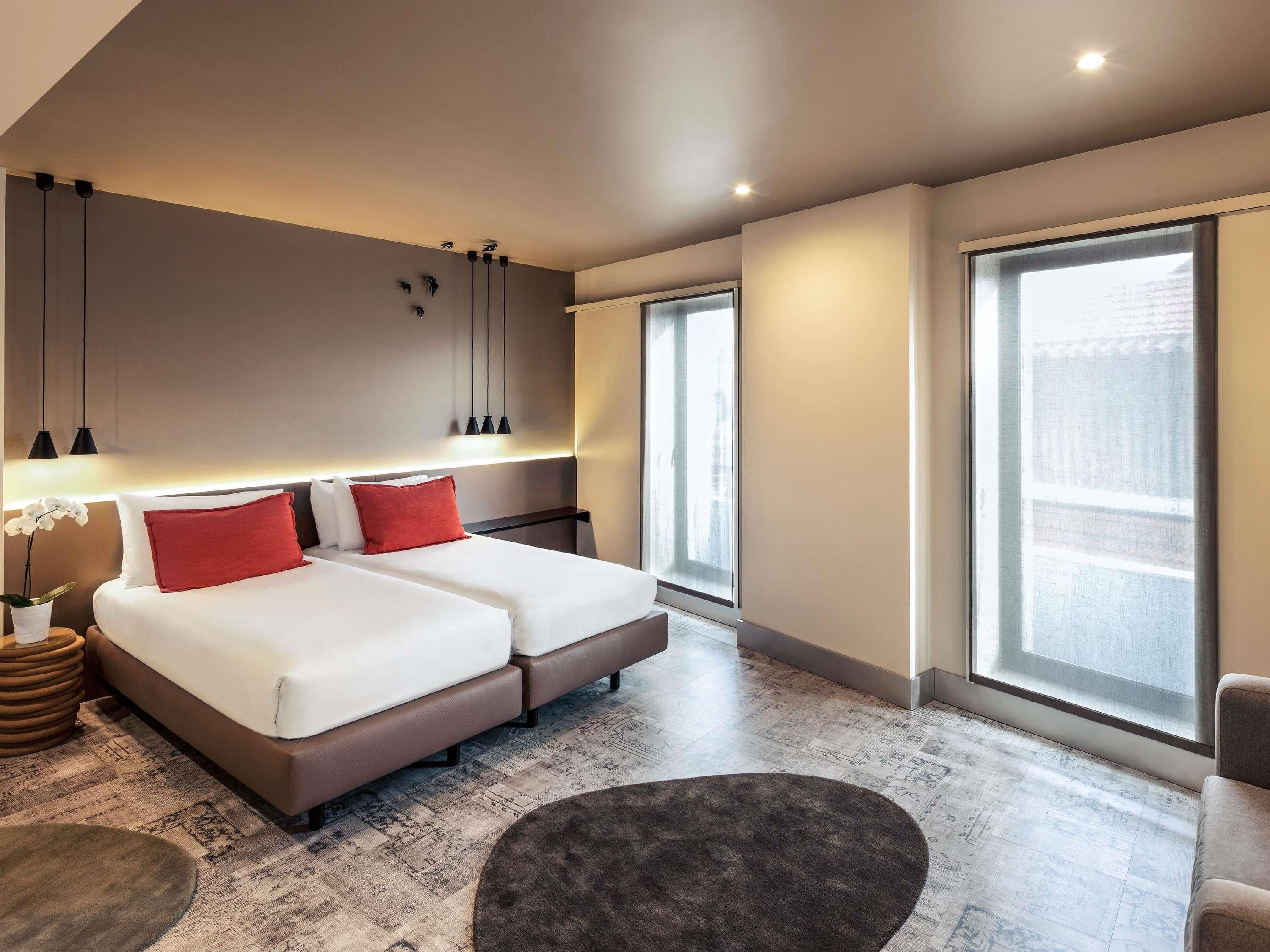 A modern hotel room with a large bed, bedside tables, a small seating area, and large windows.