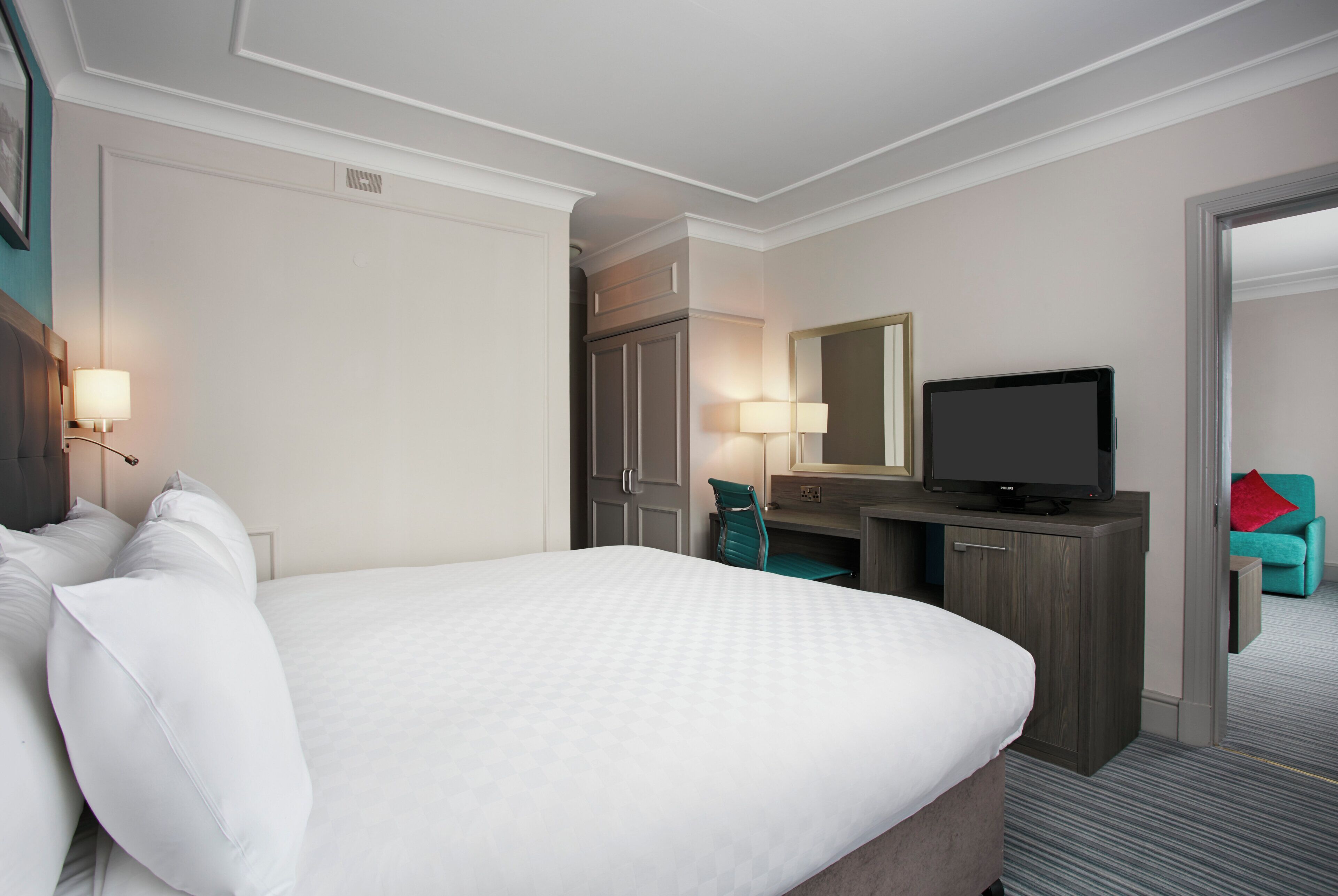 A hotel room with a large bed, a desk with a television, a chair, and a small seating area with a red pillow.