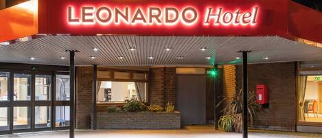The exterior of the Leonardo Hotel with a prominent illuminated sign, a brick facade, and a covered entrance area.