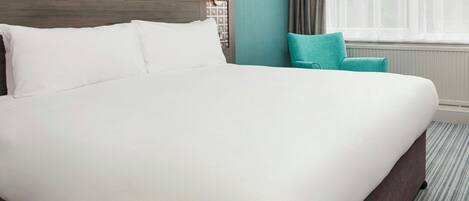 Una habitación de hotel con una cama grande, una mesita de noche con lámpara, una ventana con cortinas y una silla color turquesa.