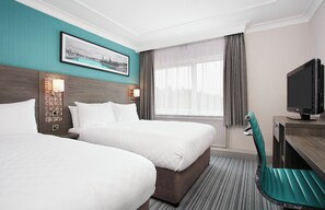Quarto de hotel com duas camas, televisão, escrivaninha e janela com cortinas.