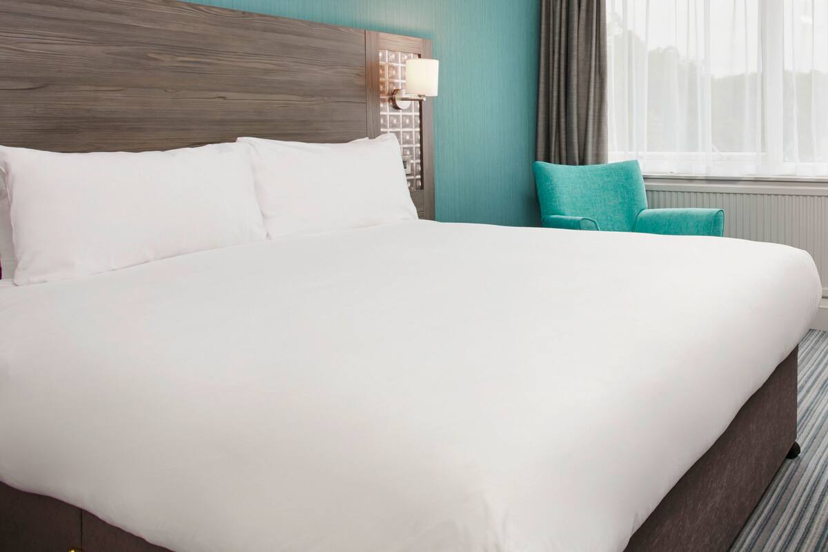 A neatly made bed with white linens, a wooden headboard, a teal accent pillow, and a lamp on a bedside table.