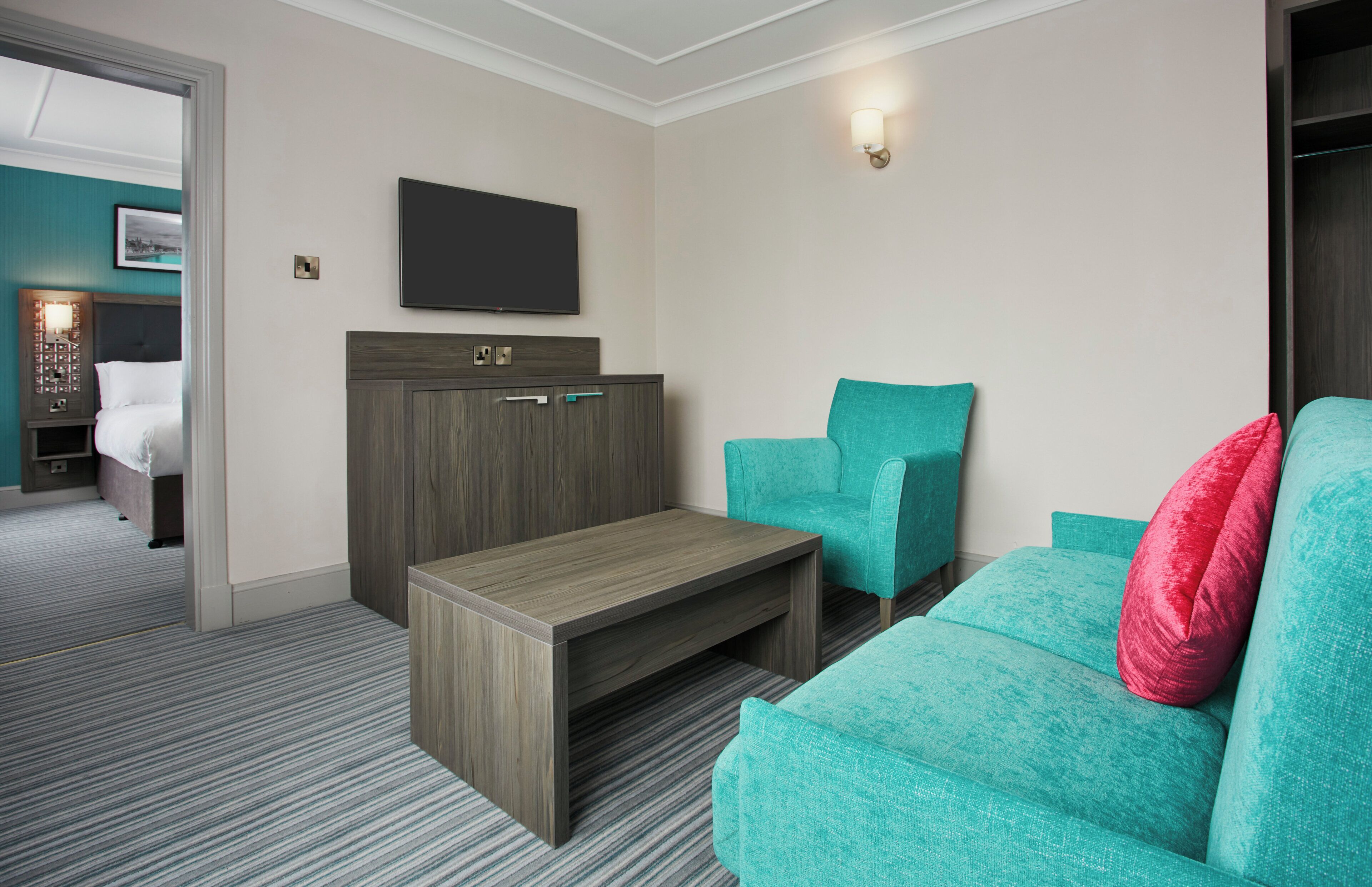 Habitación de hotel con cama, escritorio, sillón verde azulado, almohada roja y televisor montado en la pared.