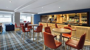 A hotel bar area with round tables and red chairs, a bar counter with a variety of bottles, and a plaid carpet. - Leonardo Hotel Inverness (Inverness)