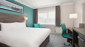 A hotel room with a large bed, a desk with a chair, a window with curtains, a wall-mounted lamp, and a framed picture on the wall.