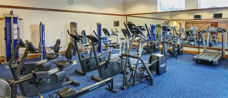 A gym with various cardio and strength training equipment, a blue carpet, and large windows.